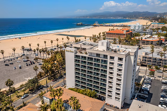 Aerial view of the Brookdale Ocean House building near a sandy beach with palm trees, parked cars, and a pier extending into the ocean under a clear blue sky.