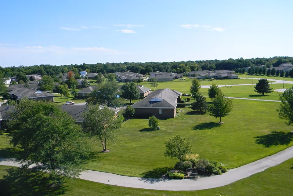 Aerial view of a senior living community with single-story buildings surrounded by well-maintained green lawns, trees, and walking paths under a clear blue sky.