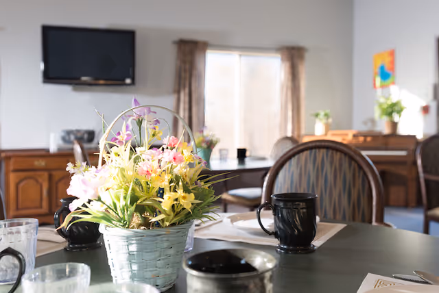 A dining area in a senior living facility with a table set with black mugs, glasses, and placemats. A basket of colorful artificial flowers is in the center of the table. In the background, there are wooden chairs, a window with curtains, a wall-mounted TV, and some potted plants on furniture.