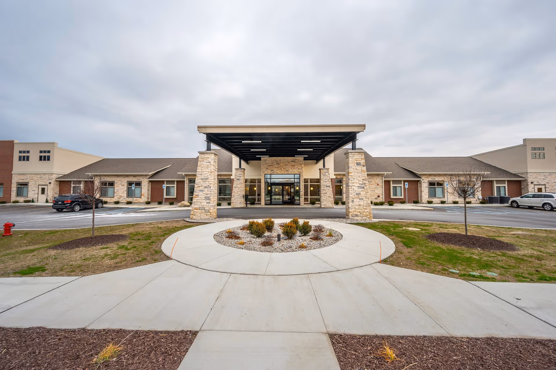 Front entrance of a senior living facility with stone pillars, a covered porte-cochère and a circular driveway under a cloudy sky.
