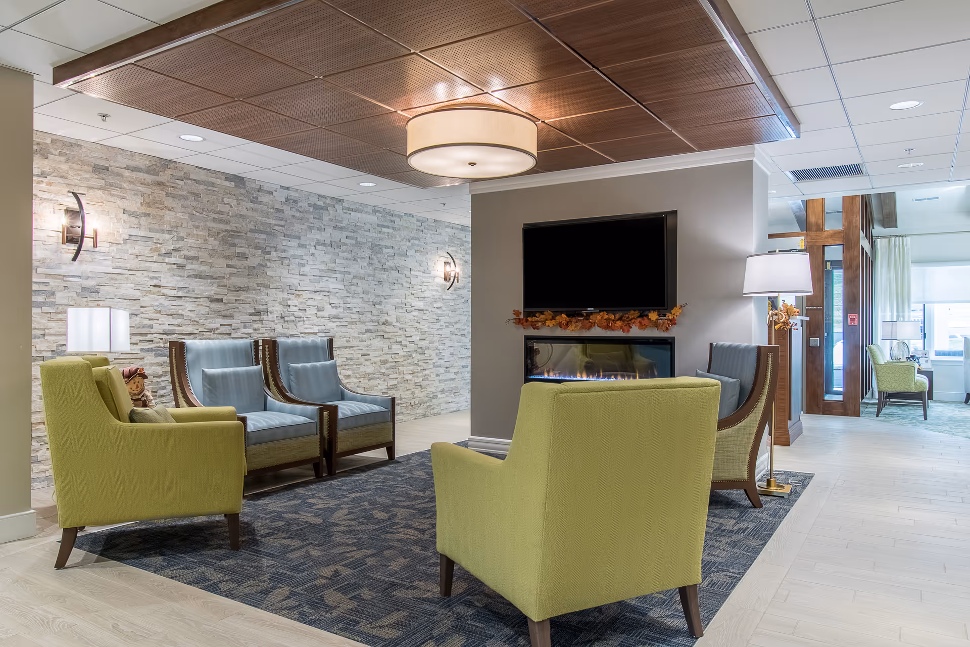 A modern lounge with green and blue armchairs arranged around a fireplace and wall-mounted TV in a bright common area.