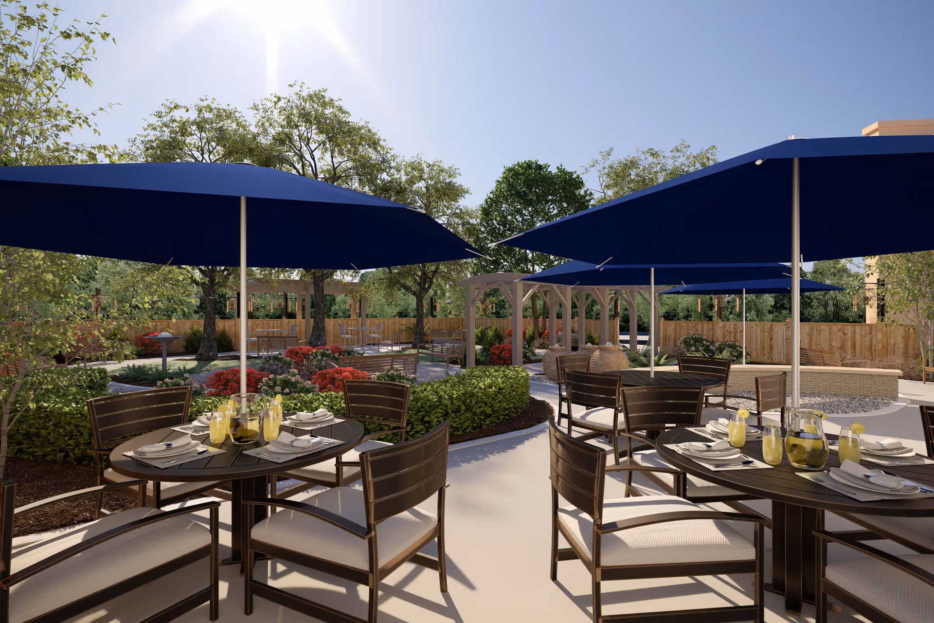 Outdoor patio with round dining tables set with drinks and plates under navy blue umbrellas, surrounded by trees, flowers, and a pergola.
