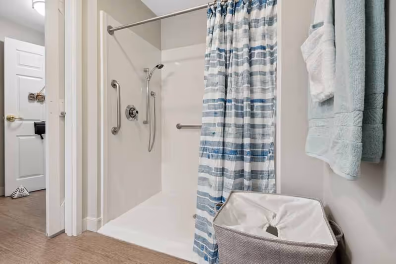 Accessible bathroom shower with a blue-striped curtain, grab bars, a handheld showerhead, towels, and a laundry basket.
