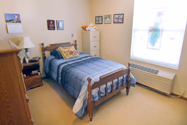 A small bedroom with a wooden single bed covered with a blue plaid comforter and a decorative pillow. There is a wooden nightstand with a lamp and some items on it next to the bed. A wooden dresser is partially visible on the left side. On the right side, there is a white filing cabinet with some items on top, and a window with blinds letting in natural light. The walls have a few framed pictures and plaques.