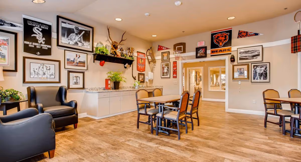 A cozy common area with wooden flooring, several tables and chairs, and two black armchairs. The walls are decorated with framed sports memorabilia, including Chicago White Sox and Chicago Bears banners and photographs. There are plants on a countertop and a small side table, and the room is well-lit with recessed ceiling lights.