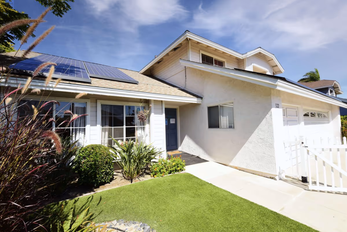 Exterior view of a residential-style building with solar panels on the roof, a blue front door, large windows, and a well-maintained front yard with green grass and various plants under a partly cloudy sky.