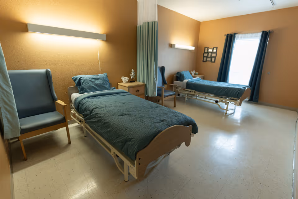 Shared nursing facility bedroom with two hospital-style beds, chairs, and a bedside table.