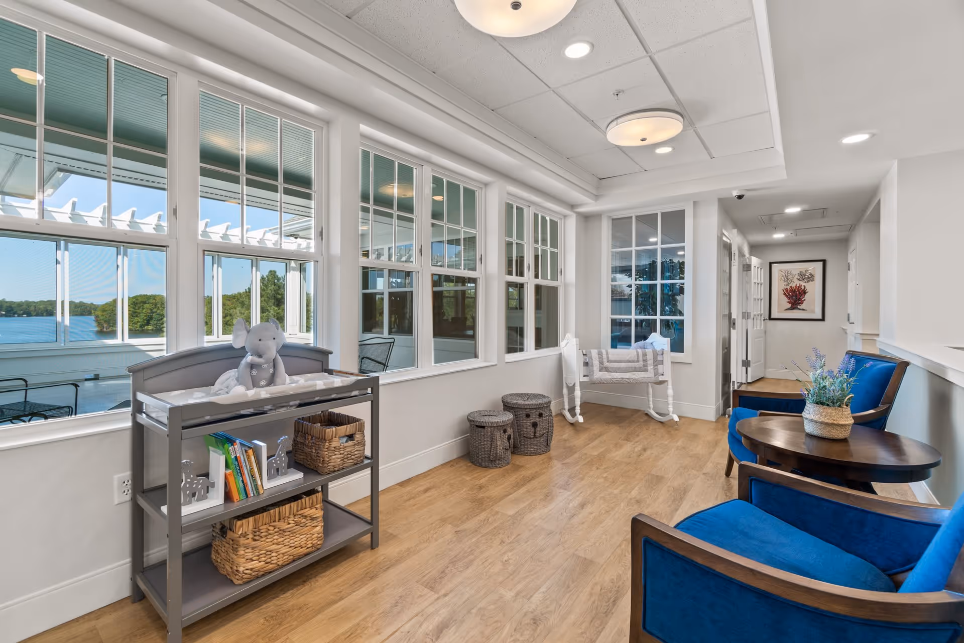 A bright and airy sitting area with large windows overlooking a lake. The room features two blue upholstered chairs with a small round wooden table between them, a gray changing table with baskets and children's books, two woven storage baskets, and a white rocking cradle with a quilt. The floor is wood, and the walls are painted white with a framed coral artwork at the end of the hallway.