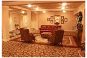 A cozy living room area with patterned carpet, a red leather sofa, two upholstered armchairs, a wooden coffee table, and framed artwork on the walls. The room is softly lit with recessed ceiling lights and has a warm, inviting atmosphere.