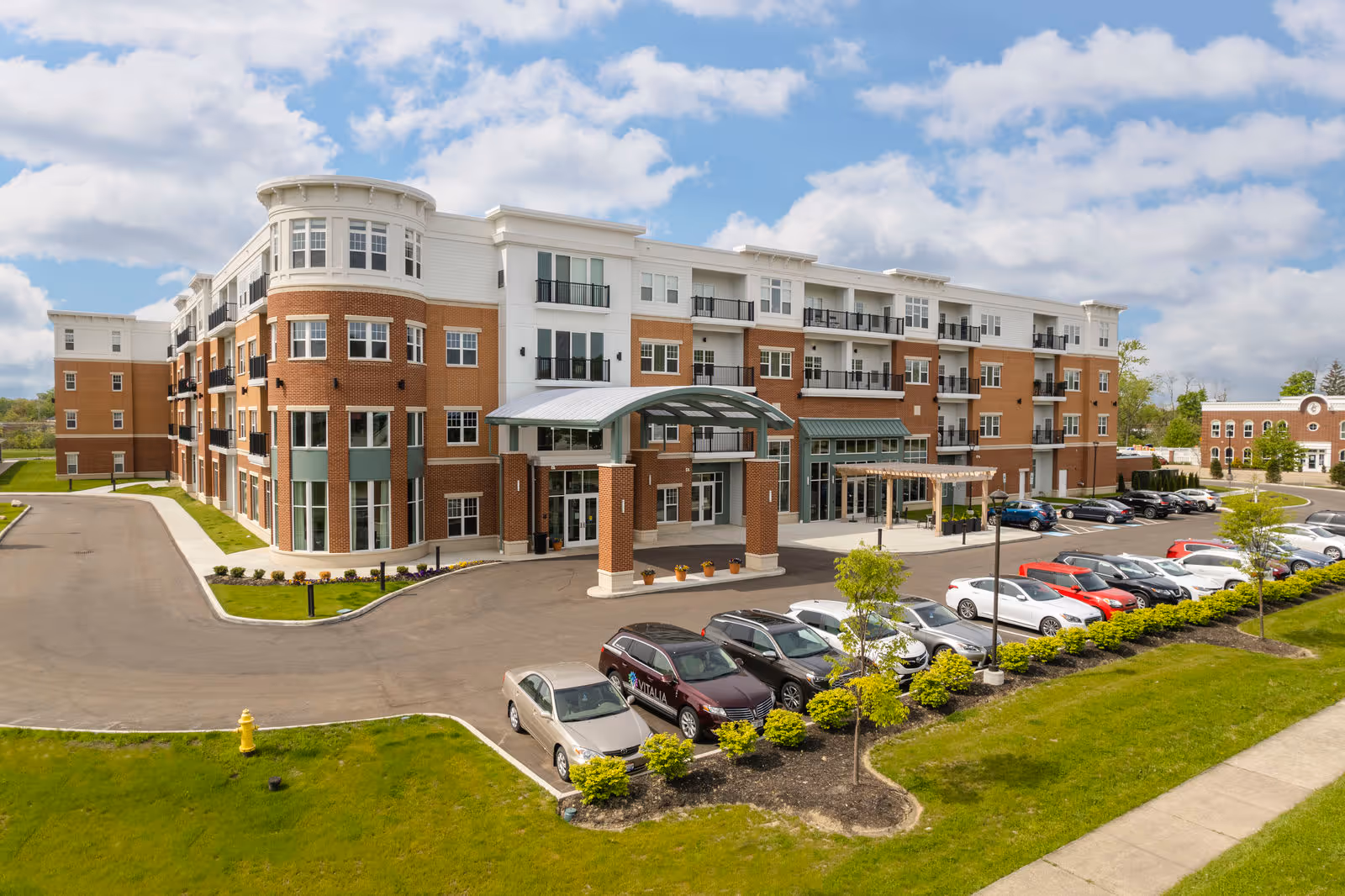 Multi-story brick and white senior living building with a covered main entrance, balconies, and a front parking lot.