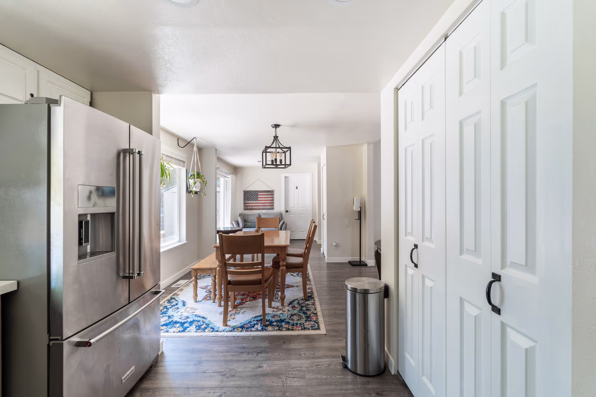 Interior view of a dining area in a senior living facility with a wooden dining table and chairs on a colorful rug. To the left is a stainless steel refrigerator, and to the right are white paneled closet doors. The room has light-colored walls and flooring, with a hanging light fixture above the table and a window with hanging plants.