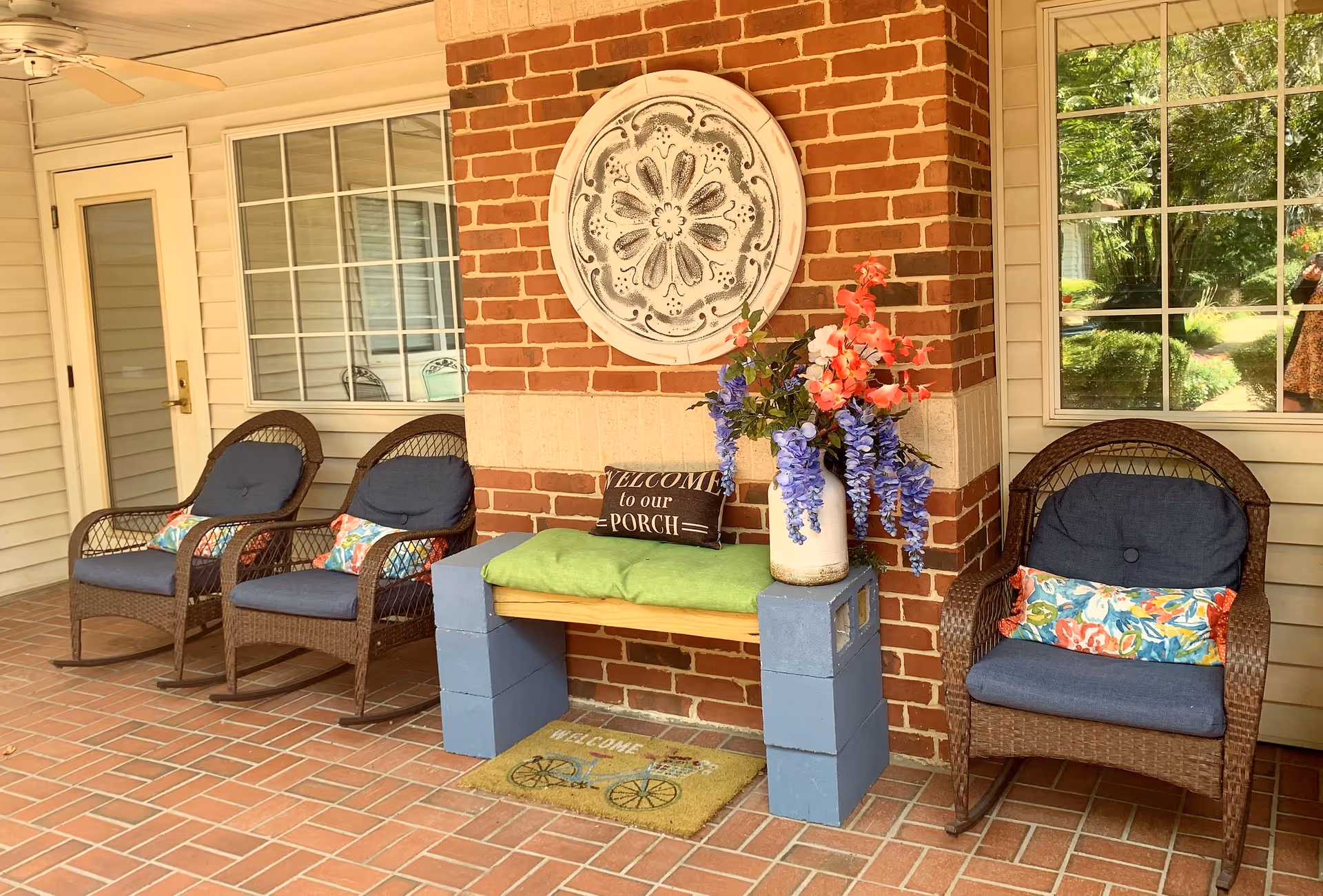 Covered porch seating area with wicker chairs, a small bench, and decorative flowers against a brick column.