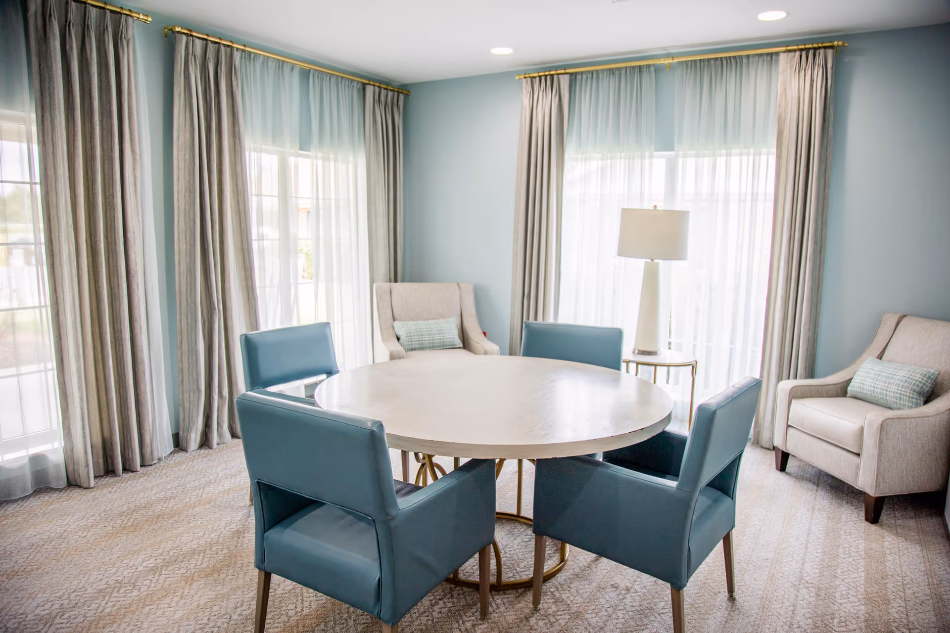 Bright seating area with a round table surrounded by teal chairs and armchairs near large windows with floor-length curtains.