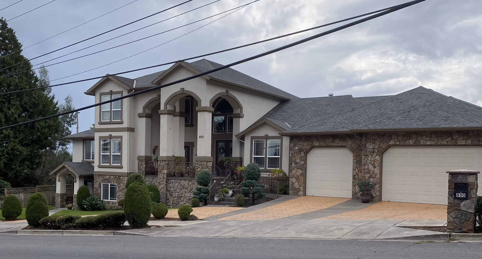 Exterior view of a large two-story residential building with a stone and stucco facade, multiple windows, a double garage with beige doors, a driveway, and well-maintained landscaping including trimmed bushes and small trees. The building has an arched entrance and the number 800 is visible near the front door and on the mailbox.