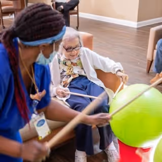 An elderly woman sitting in a chair playing a game with a caregiver who is holding drumsticks and a large green exercise ball in front of them in a common area with wooden flooring and chairs.