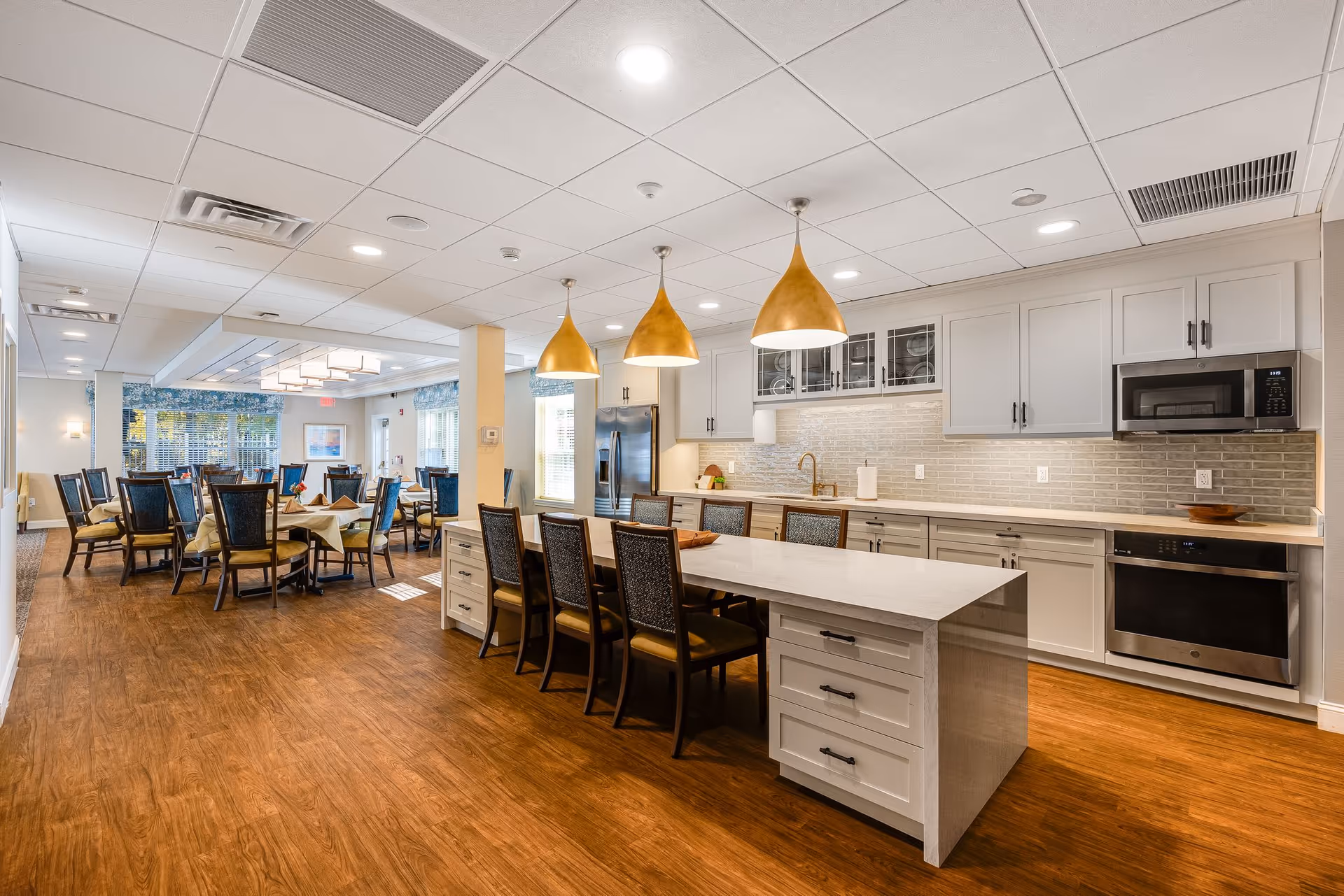 A spacious and well-lit dining area combined with a kitchen in a senior living facility. The kitchen features white cabinets, a long island with seating for six, modern appliances including a microwave and oven, and three large pendant lights hanging above the island. The dining area has multiple tables with chairs arranged neatly, and large windows allowing natural light to fill the space.