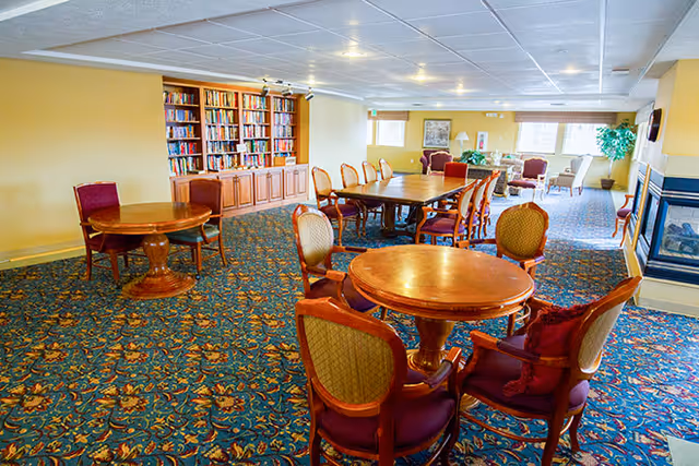 A spacious common area with multiple wooden tables and upholstered chairs arranged on a patterned carpet. There is a large bookshelf filled with books against one wall, a fireplace on the right, and several windows letting in natural light. The room has a cozy and inviting atmosphere.
