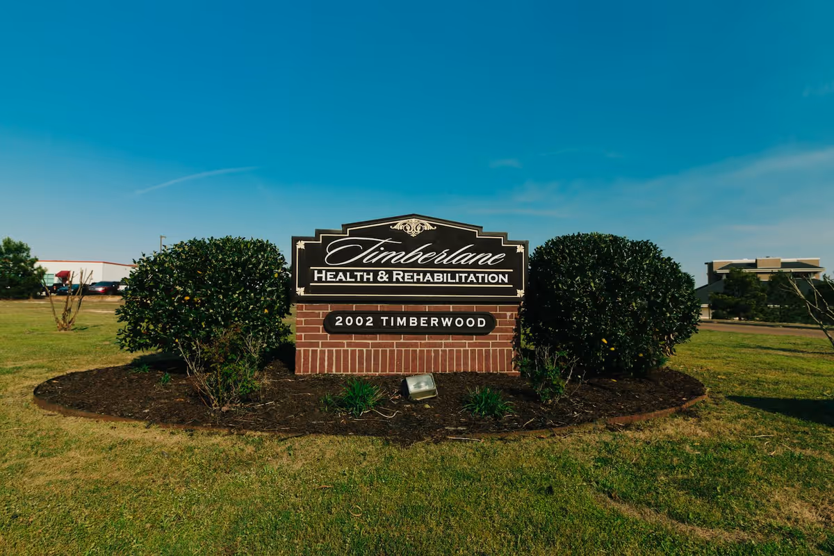 Outdoor brick sign reading 'Timberlane Health & Rehabilitation' with '2002 Timberwood' on a landscaped lawn flanked by bushes and buildings in the background.