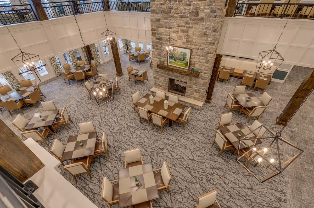 A spacious dining room with multiple square tables and beige cushioned chairs arranged neatly. The room features a large stone fireplace with a painting above it, modern hanging light fixtures, large windows with patterned curtains, and a carpeted floor with a branch-like design.