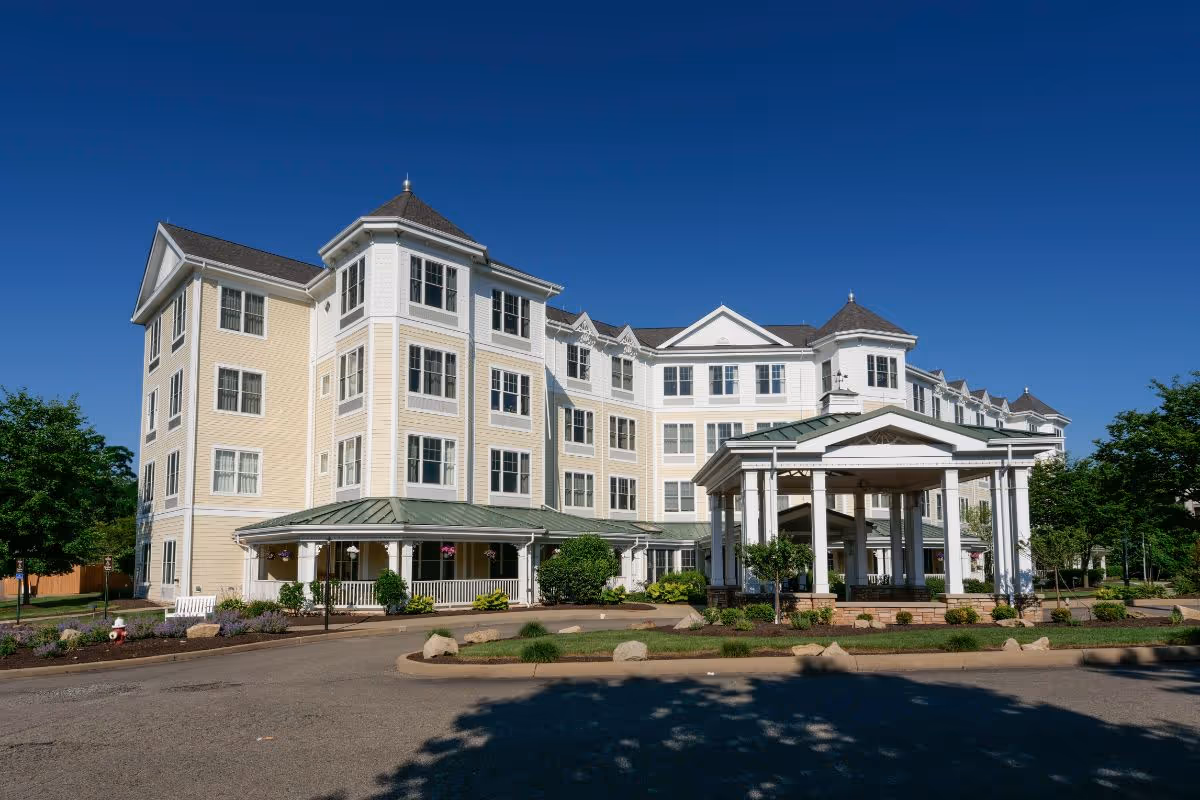 Exterior view of a large, multi-story senior living facility building with beige and white siding, multiple windows, and a covered entrance area. The building is surrounded by landscaped greenery and a clear blue sky is visible above.