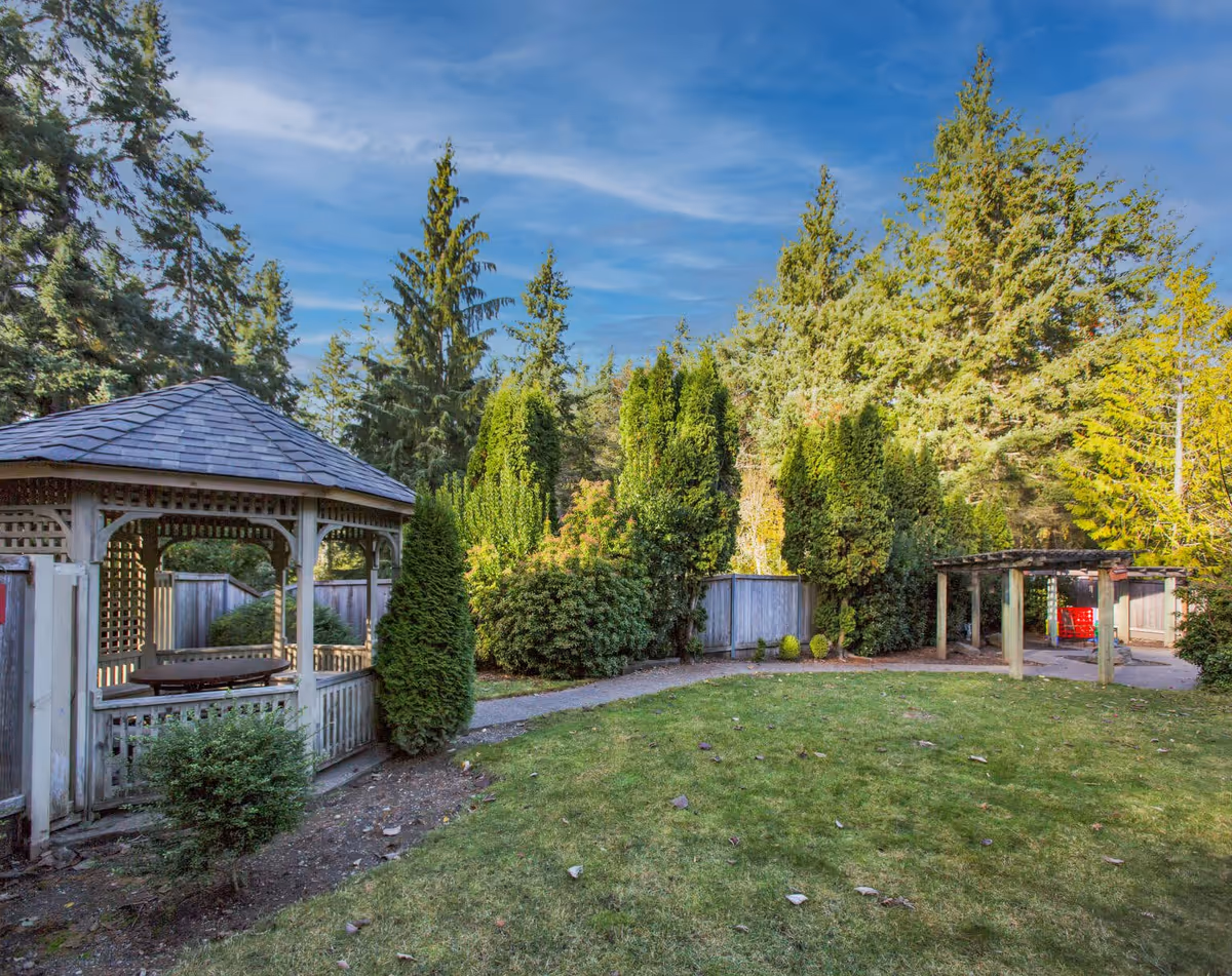 A peaceful outdoor garden area at Greenlake Senior Living Gig Harbor featuring a wooden gazebo with a shingled roof on the left, surrounded by green shrubs and tall trees. A pathway runs through the garden, and there is a wooden pergola structure on the right side. The sky is clear with some wispy clouds.