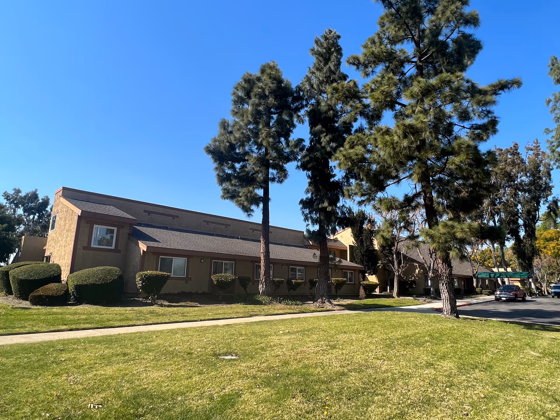Exterior view of a single-story building with beige walls and brown trim, surrounded by tall pine trees and neatly trimmed bushes, under a clear blue sky. There is a green lawn in the foreground and a driveway with parked cars on the right side.