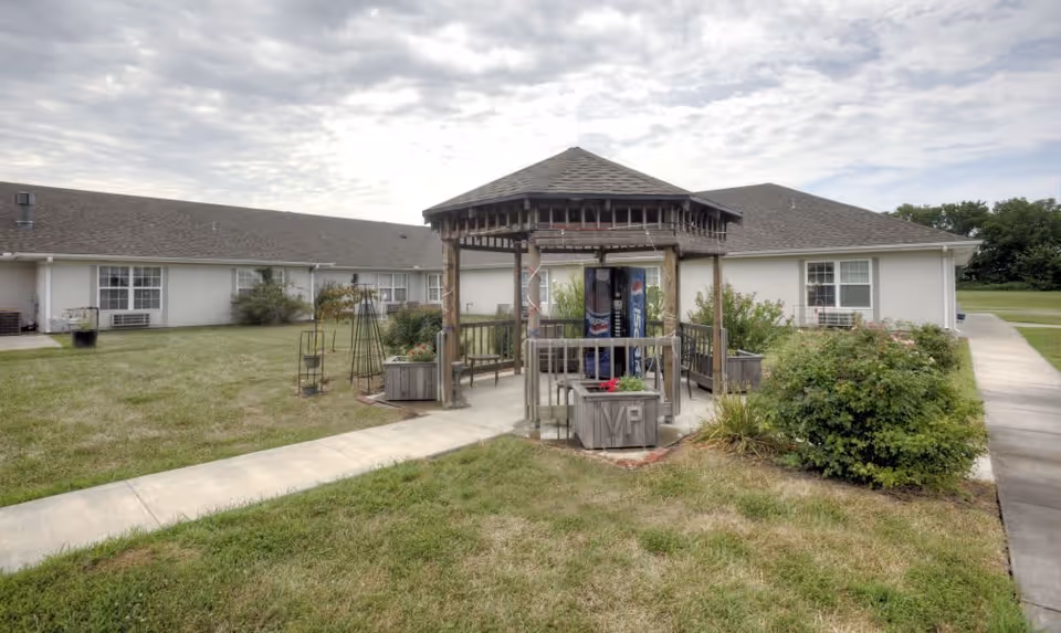 Outdoor view of a senior living facility with a wooden gazebo in the center surrounded by grass and bushes. The building is single-story with white walls and multiple windows. A vending machine is visible inside the gazebo. Concrete walkways lead to and from the gazebo.