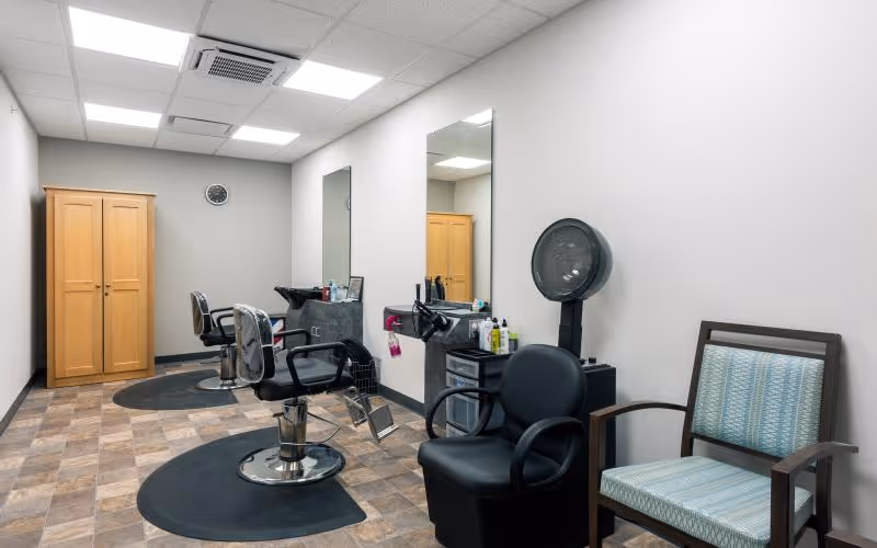 Interior view of a hair salon area with two black salon chairs on black mats, a hair dryer chair, a wooden cabinet, two large mirrors, and a couple of chairs with patterned upholstery against the wall.