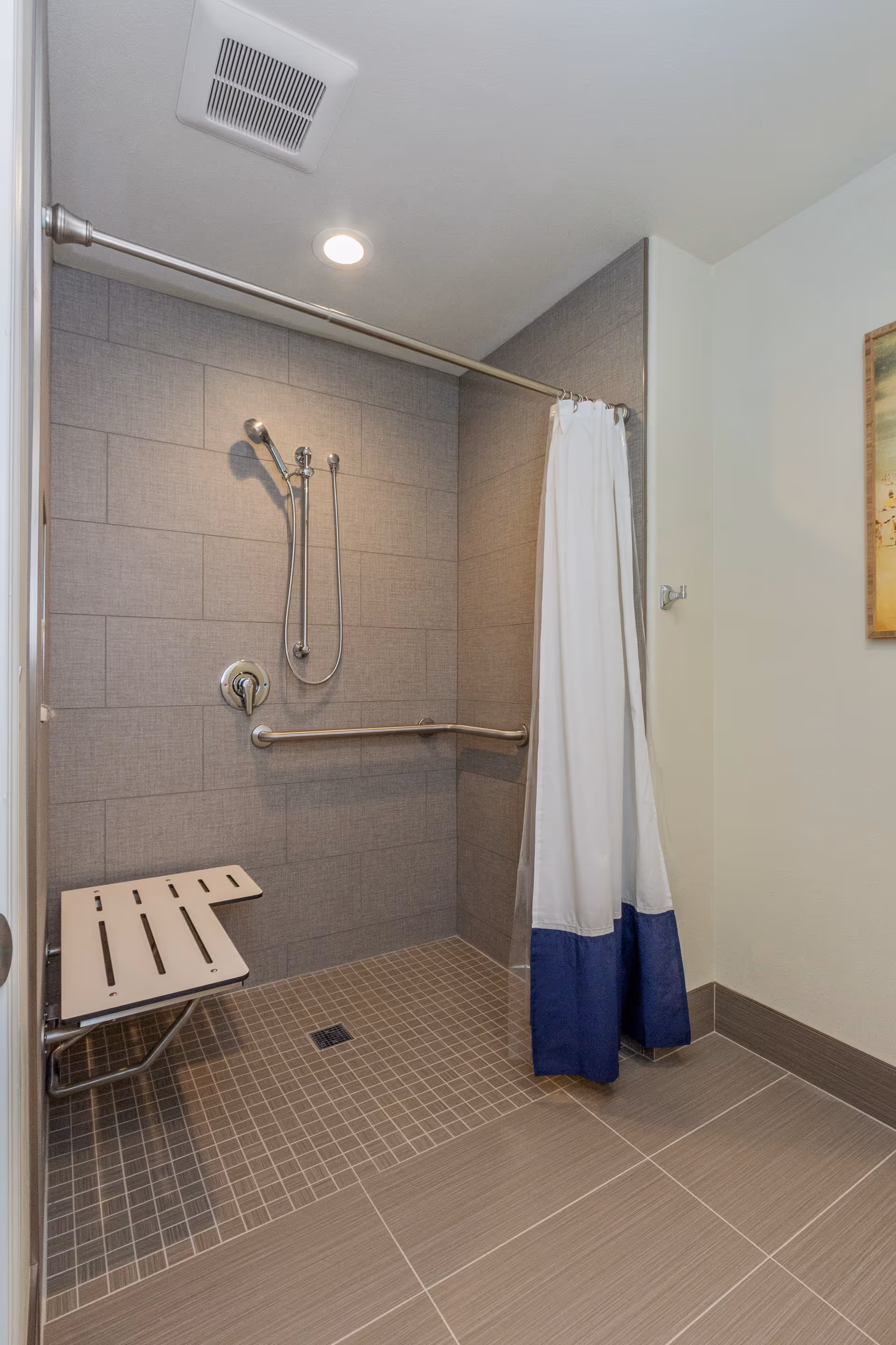 A modern accessible shower area with gray tiled walls and floor, a white and blue shower curtain, a foldable shower seat, a handheld showerhead, and a horizontal grab bar.