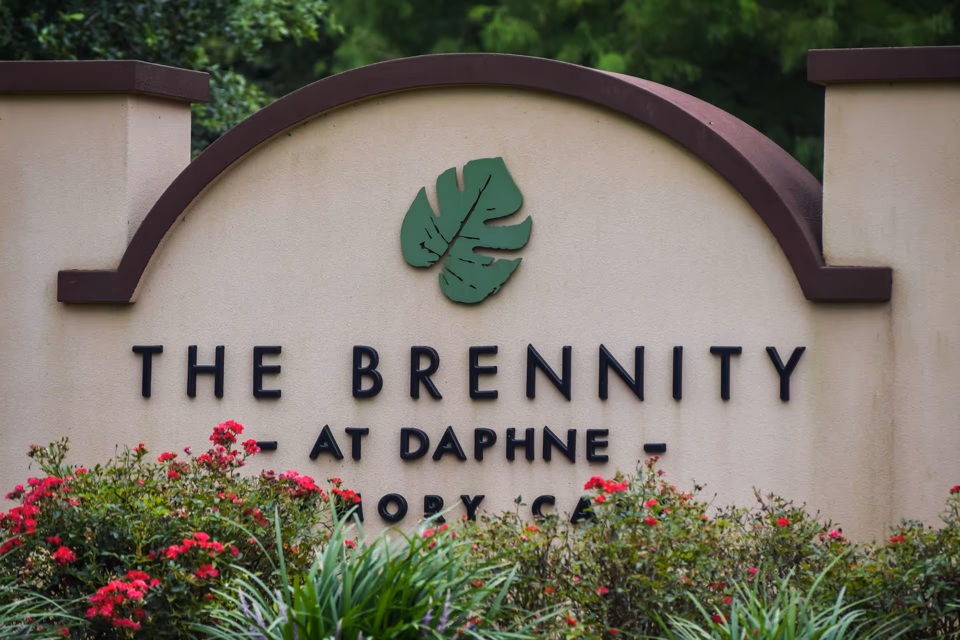 A beige and brown entrance sign for The Brennity at Daphne Assisted Living & Memory Care, surrounded by green plants and red flowers.