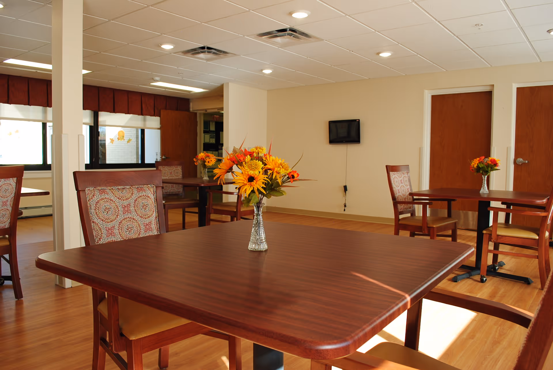 Bright dining room with wooden tables and chairs, each table topped with a small vase of flowers.