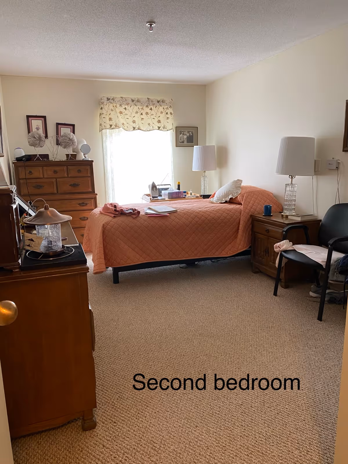 A tidy second bedroom with a single bed covered by a coral quilt, wooden dressers and nightstands, lamps, a chair, and a window with a floral valance.