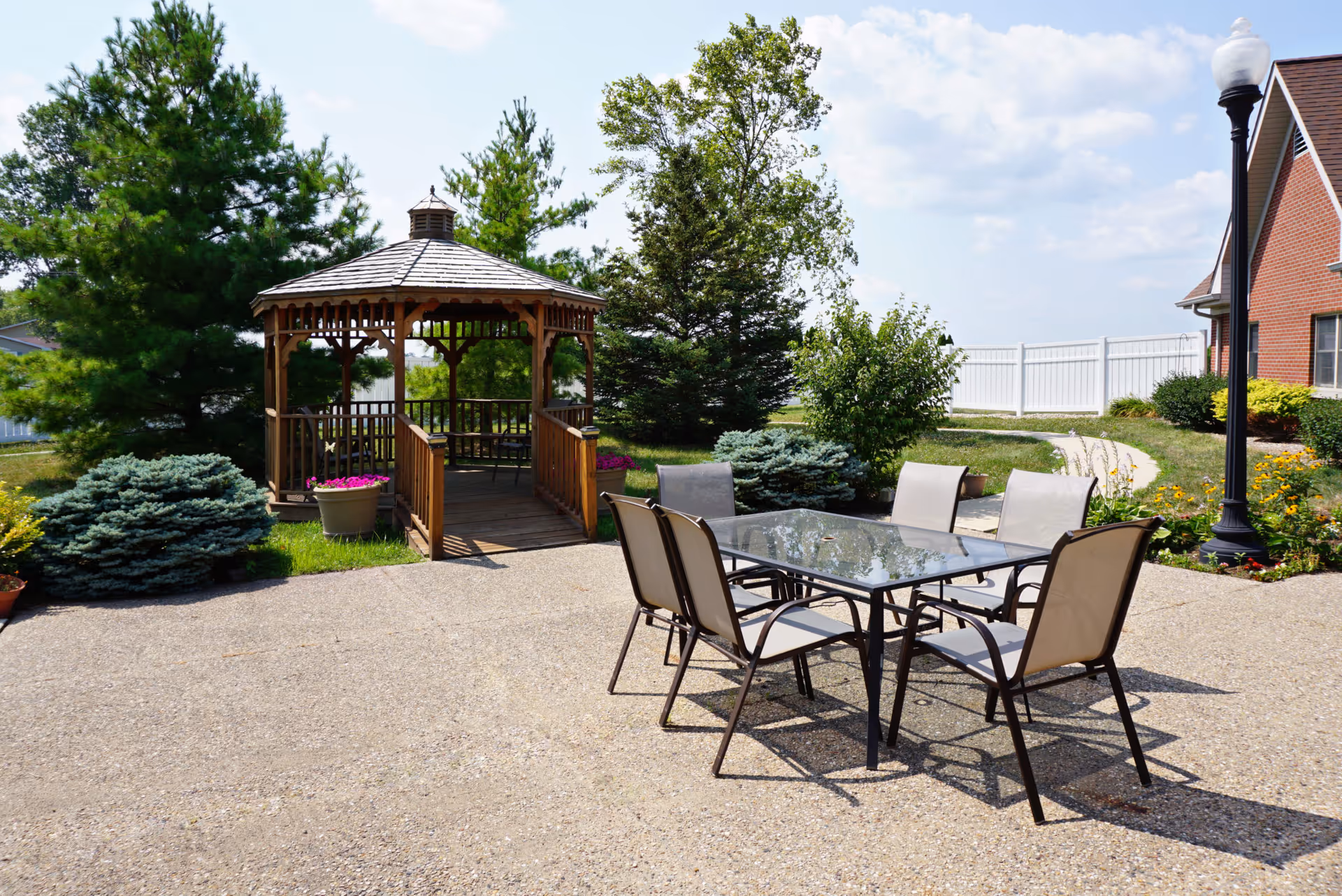 Outdoor patio with a glass-top table and chairs in front of a wooden gazebo and landscaped grounds.