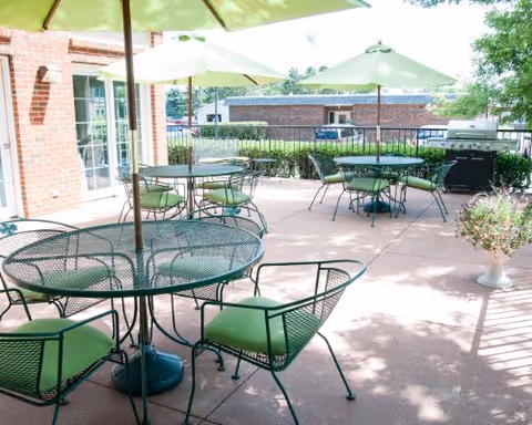 Outdoor patio with green metal tables and chairs under umbrellas next to a brick building and a grill.