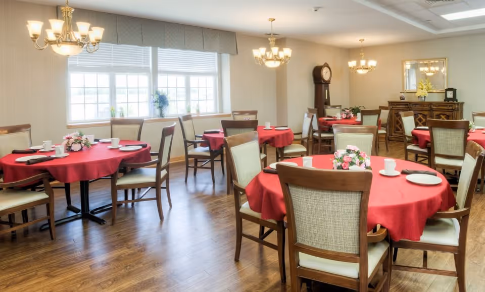 A bright dining room with several round tables covered in red tablecloths, each set with white plates, cups, and napkins. The room has wooden chairs with cushioned seats, large windows letting in natural light, chandeliers hanging from the ceiling, a grandfather clock, and a wooden sideboard with a mirror and floral arrangement.