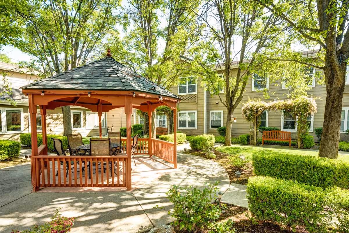 A sunny outdoor courtyard area at Vista Roseville Senior Living featuring a wooden gazebo with a shingled roof and outdoor seating including a table and chairs. The courtyard is surrounded by green bushes, trees, and a beige two-story building with multiple windows. There is also a wooden bench under an archway covered with flowering vines.