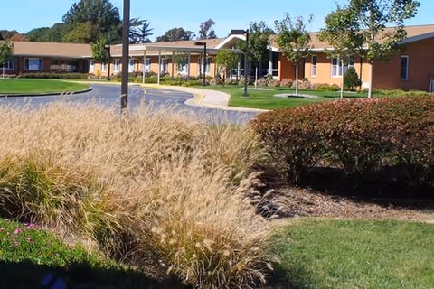 Exterior view of Commonwealth Senior Living At Eastern Shore showing a single-story brick building with a covered entrance, surrounded by well-maintained landscaping including ornamental grasses, bushes, and trees under a clear blue sky.