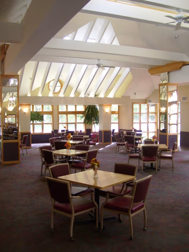 Spacious dining room with multiple square tables each surrounded by four chairs with maroon cushions. The room has large windows allowing natural light, high vaulted ceilings with white beams, ceiling fans, and decorative plants near the windows. Small flower arrangements are placed on each table.