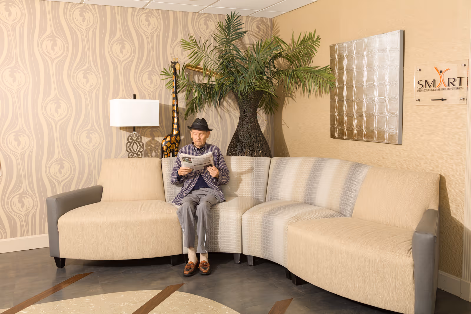 An elderly man wearing a hat and checkered shirt sits on a beige and gray sectional sofa in a lounge area, reading a newspaper. Behind him is a large decorative vase with green palm leaves, a tall giraffe sculpture, a table lamp, and a wall with patterned wallpaper. A sign on the wall reads 'SMART Specialized Medical And Rehabilitation Therapy' with an arrow pointing to the right.