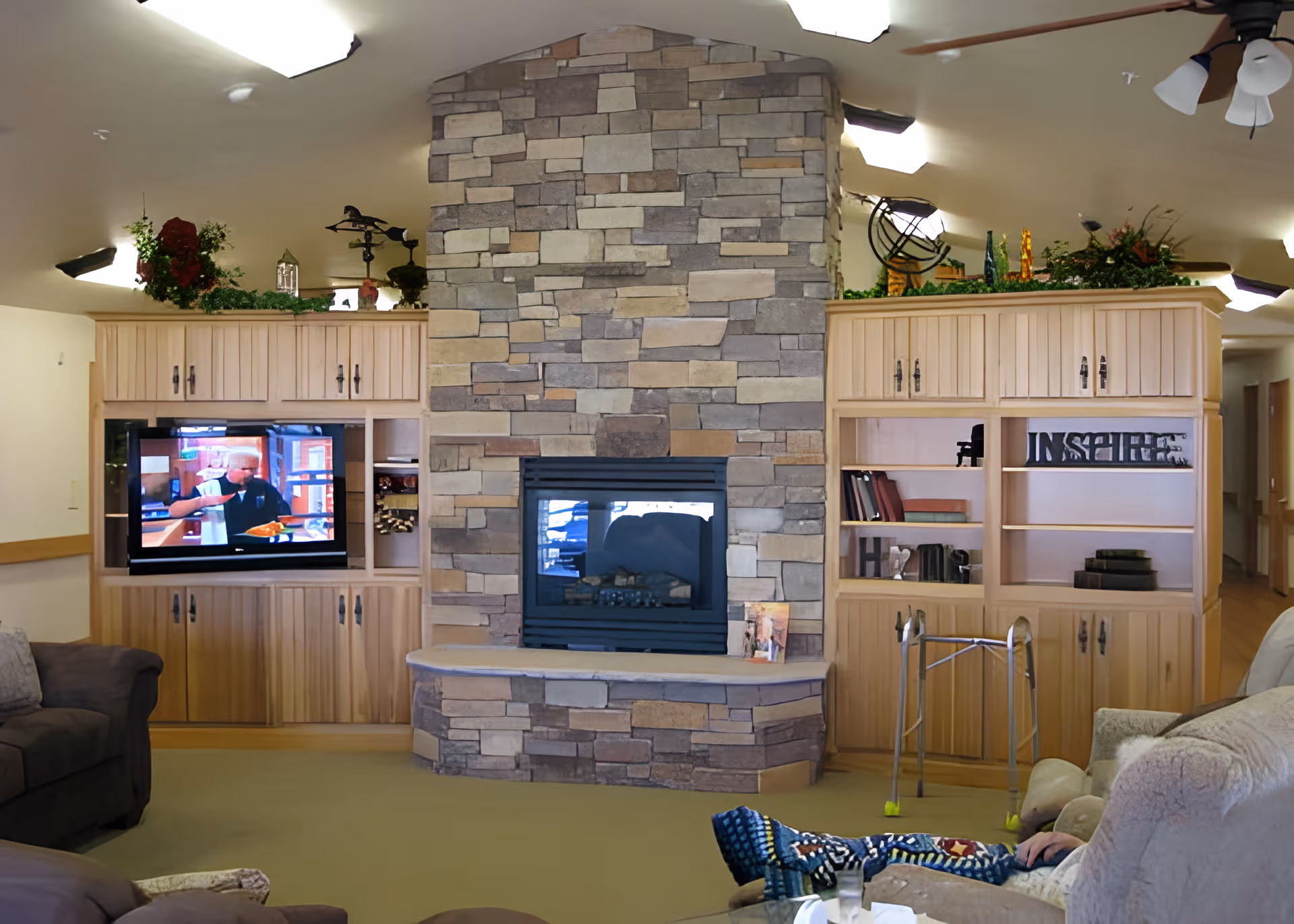 A cozy living room area featuring a stone fireplace in the center with wooden built-in cabinets on either side. The left cabinet has a television displaying a man, while the right cabinet has shelves with books and decorative items. There are comfortable sofas on both sides, a walker in front of the right cabinet, and a person partially visible sitting on the right sofa. The ceiling has recessed lighting and a ceiling fan.