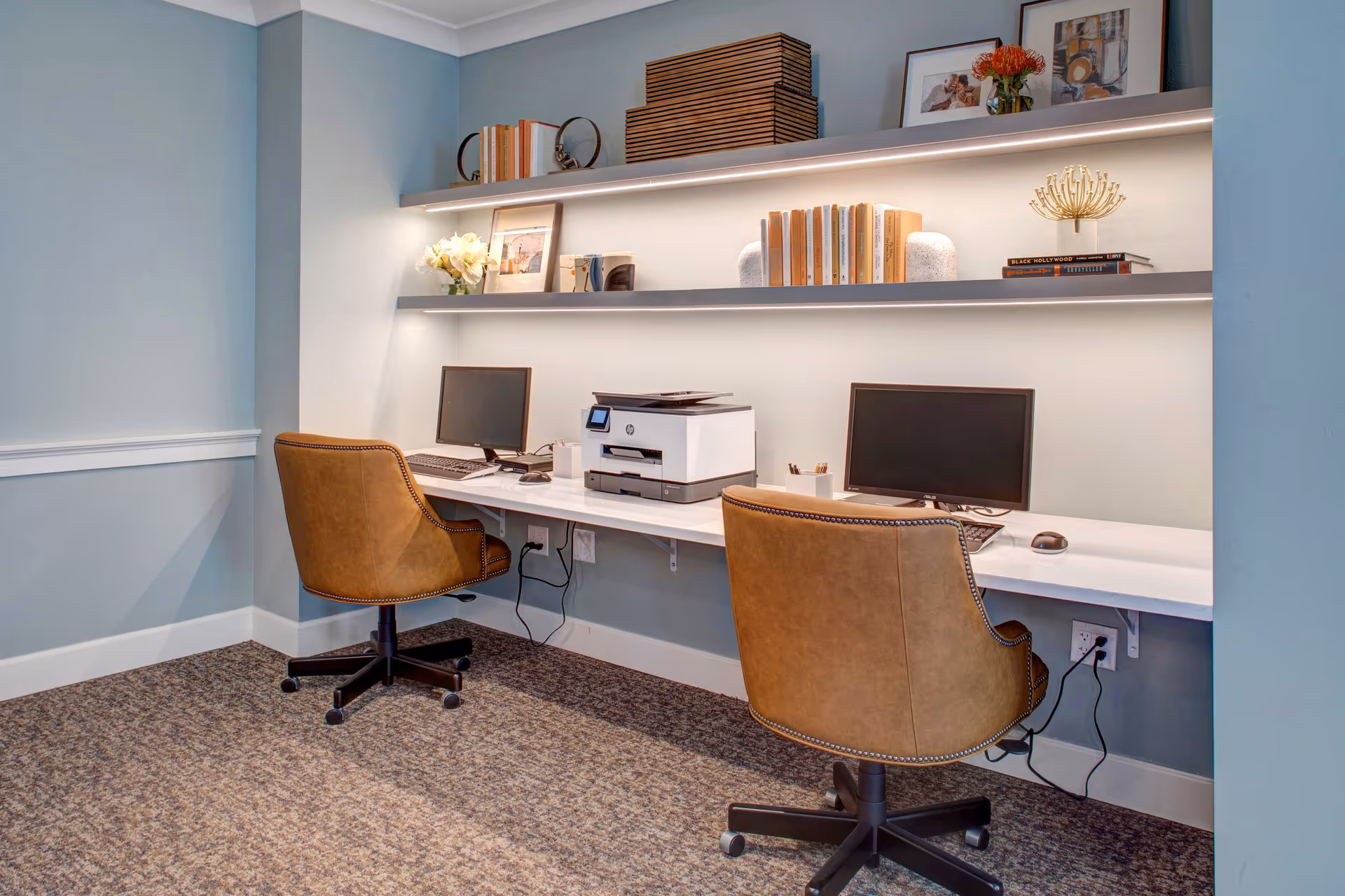 A small office area with two brown leather swivel chairs in front of a white desk mounted to the wall. On the desk are two computer monitors, keyboards, a mouse, and a printer. Above the desk are two floating shelves with books, framed pictures, decorative items, and a vase with flowers. The walls are painted light blue and the floor is carpeted.