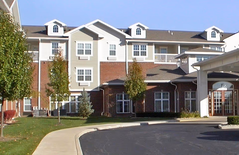 Exterior view of a multi-story senior living facility building with a combination of brick and light-colored siding, several windows, small dormer windows on the roof, and a covered entrance. There are trees and landscaped grass areas along a curved driveway in front of the building under a clear blue sky.