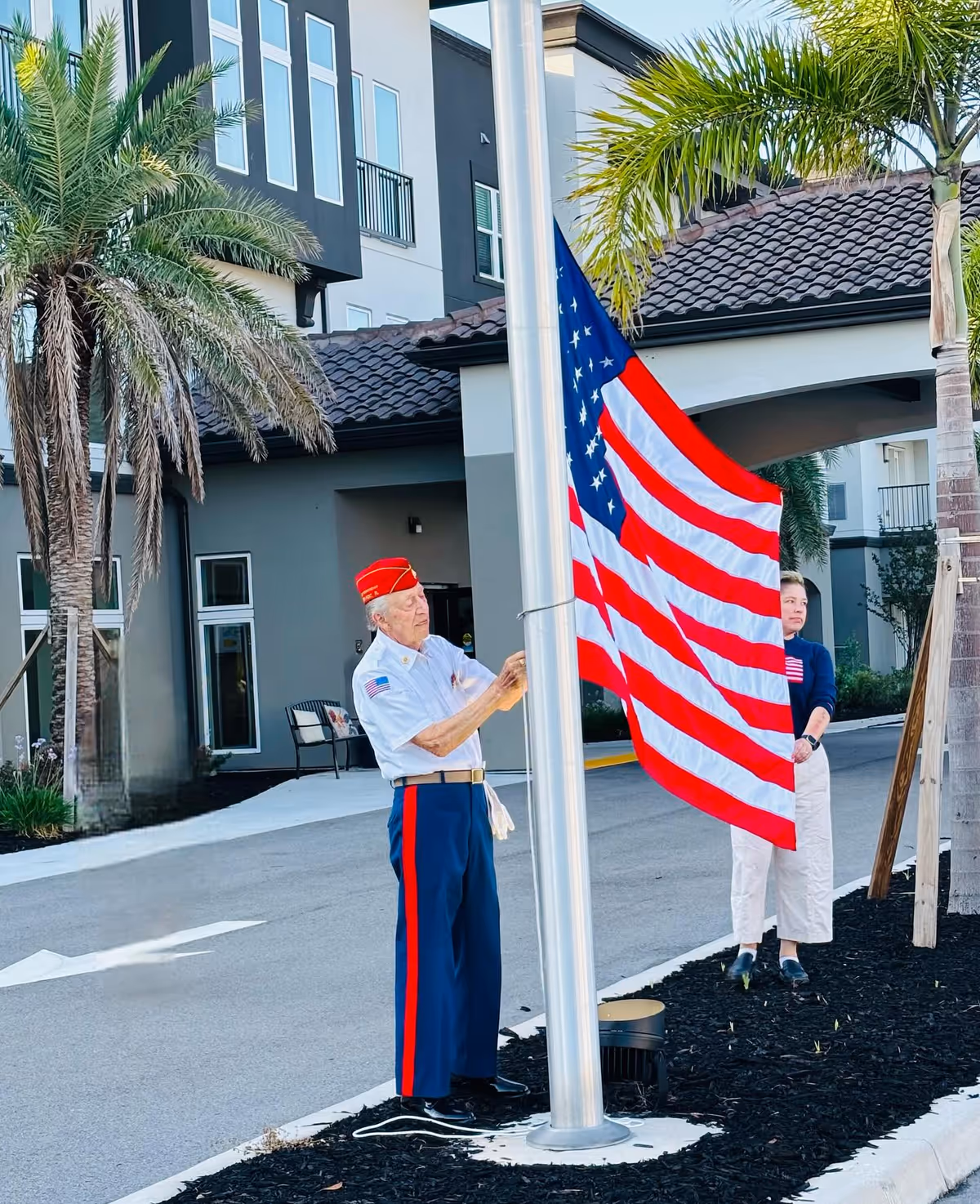 An elderly man in a white shirt and blue pants with red stripes, wearing a red hat, is raising an American flag on a flagpole outside a building. A woman stands nearby watching. The scene includes palm trees and a modern building with a tiled roof in the background.