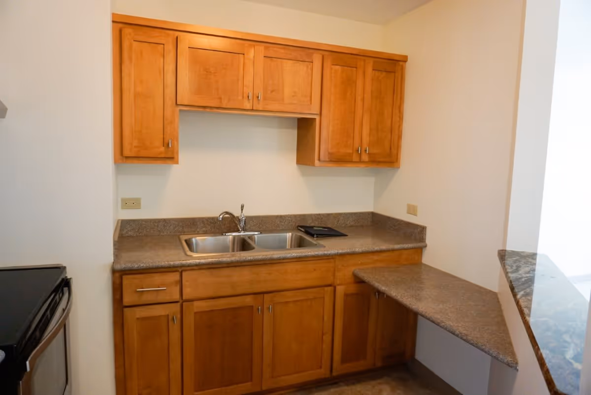 A small kitchen area with wooden cabinets, a double stainless steel sink, and a countertop that extends to form a small breakfast bar. The walls are painted white and there is an electric stove visible on the left side.