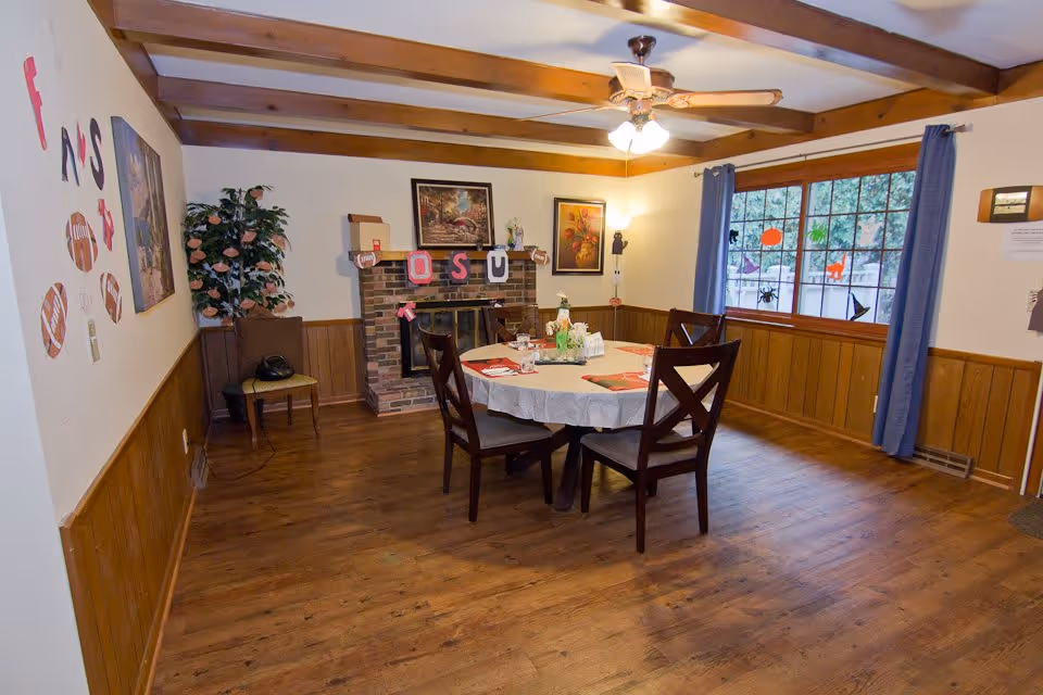 A cozy dining room with wooden floors and ceiling beams, featuring a round table covered with a white tablecloth and set with placemats, napkins, and a centerpiece. Four wooden chairs surround the table. The room has wood paneling on the lower walls and cream-colored paint above. A brick fireplace decorated with a banner and small items is on one wall, with framed artwork above it. A large window with blue curtains lets in natural light and has Halloween-themed decorations on the glass. A potted plant and a chair are placed near the fireplace.