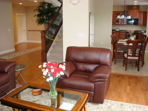 Interior view of a senior living facility showing a cozy living room with a brown leather armchair and a glass coffee table with a vase of red and white flowers. In the background, there is a dining area with a wooden dining table and chairs, and a kitchen with wooden cabinets and appliances. A staircase with a wooden railing is visible to the left.