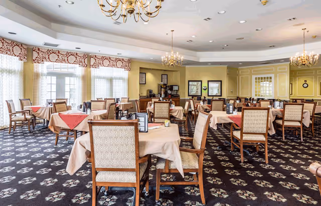A spacious dining room with multiple tables covered in beige tablecloths and red napkins. Each table is surrounded by wooden chairs with patterned upholstery. The room has large windows with sheer curtains and decorative valances, chandeliers hanging from the ceiling, and a carpet with a floral pattern. There is a beverage station against the far wall with coffee machines and framed pictures on the walls.