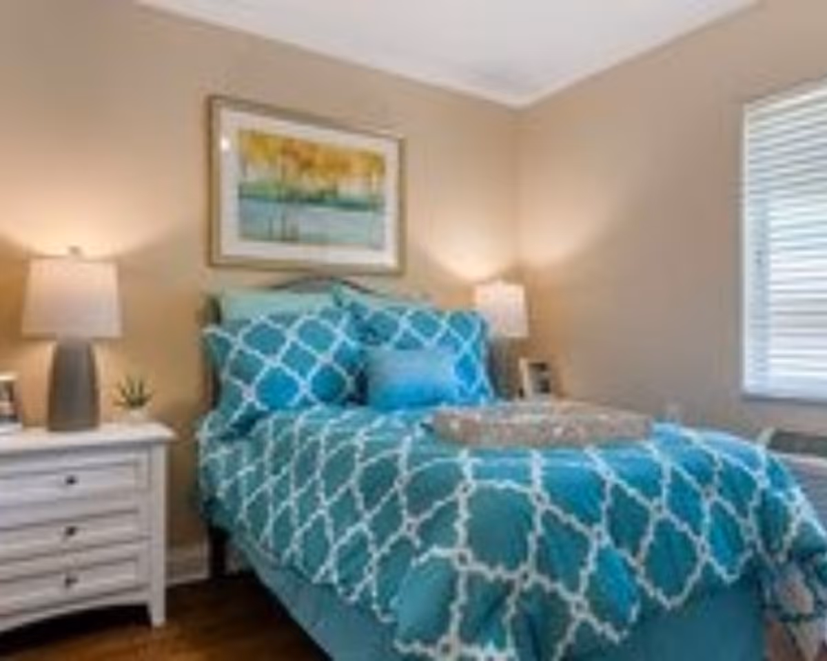 A cozy bedroom with a bed dressed in teal and white patterned bedding, two matching pillows, and a small decorative pillow. There are two bedside tables, each with a lamp and small decor items. A framed artwork hangs above the bed, and a window with blinds is on the right wall.