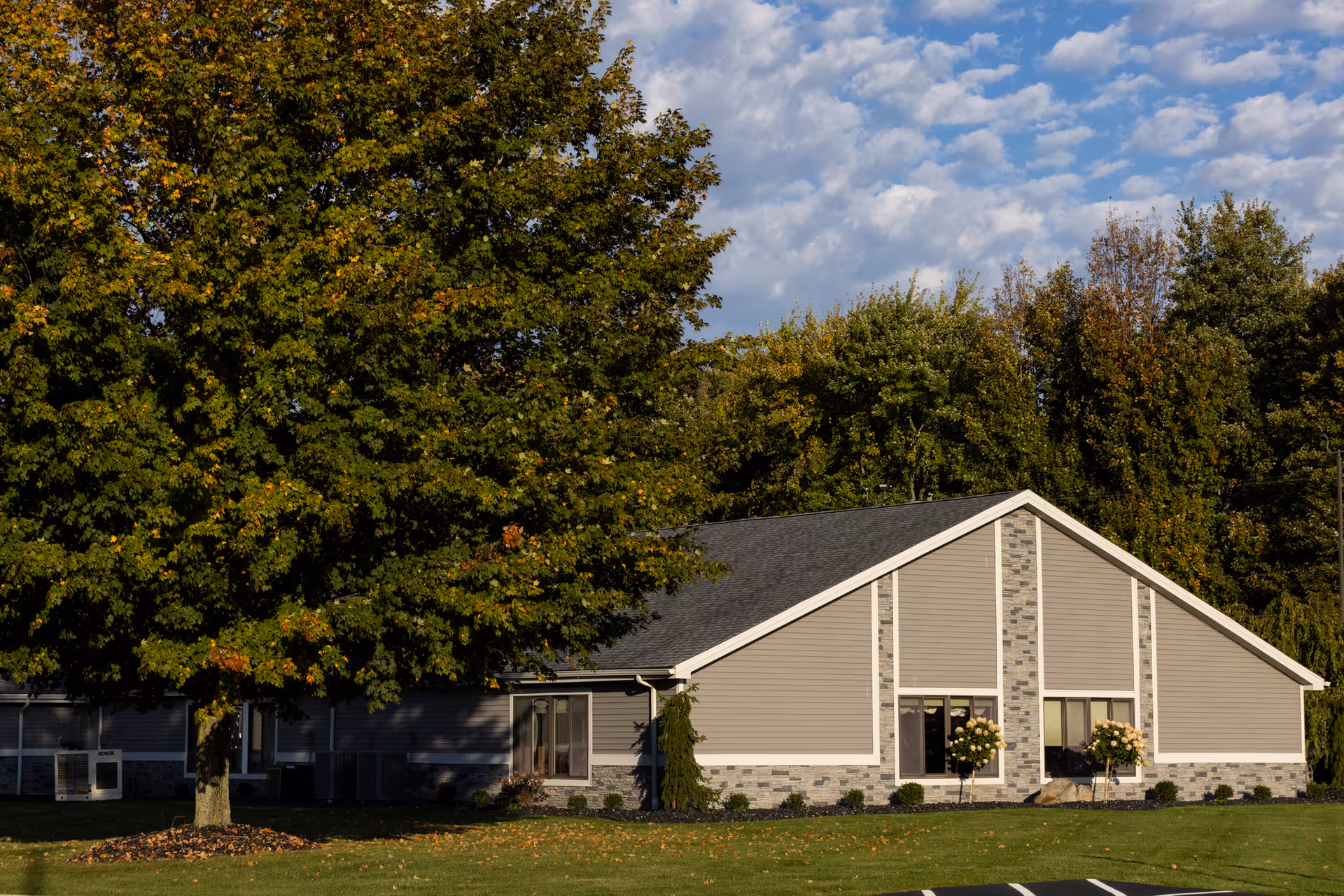 Exterior view of a single-story building with gray siding and stone accents, surrounded by green grass and trees under a partly cloudy sky.