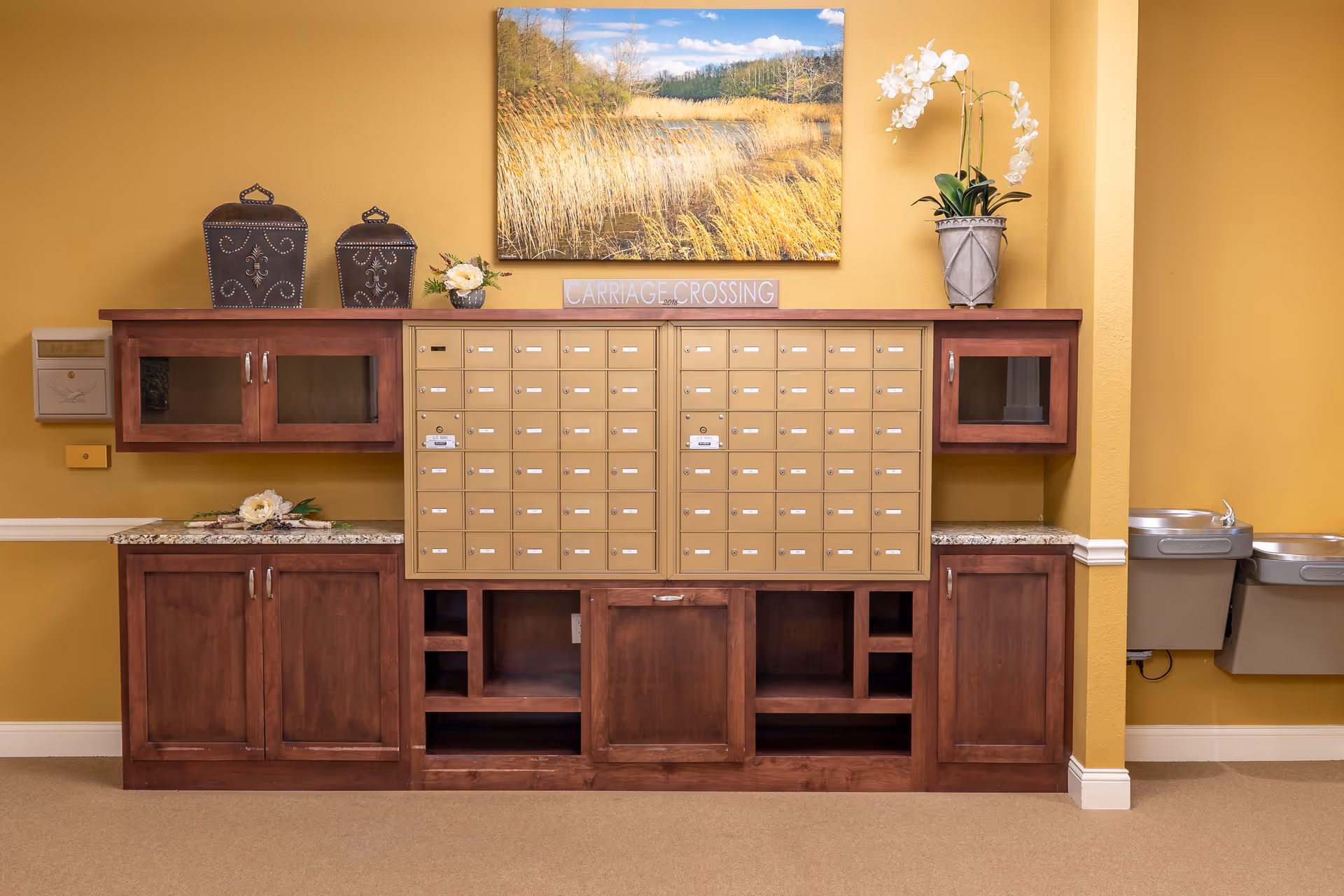 A mailroom area with multiple beige mailboxes set into a wooden cabinet with dark wood finish. Above the mailboxes is a landscape painting of a field with tall grasses and trees under a blue sky. Decorative items including two dark metal containers, a small flower arrangement, and a potted white orchid are placed on top of the cabinet. To the right, two silver water fountains are mounted on the wall. The walls are painted yellow and the floor is carpeted.